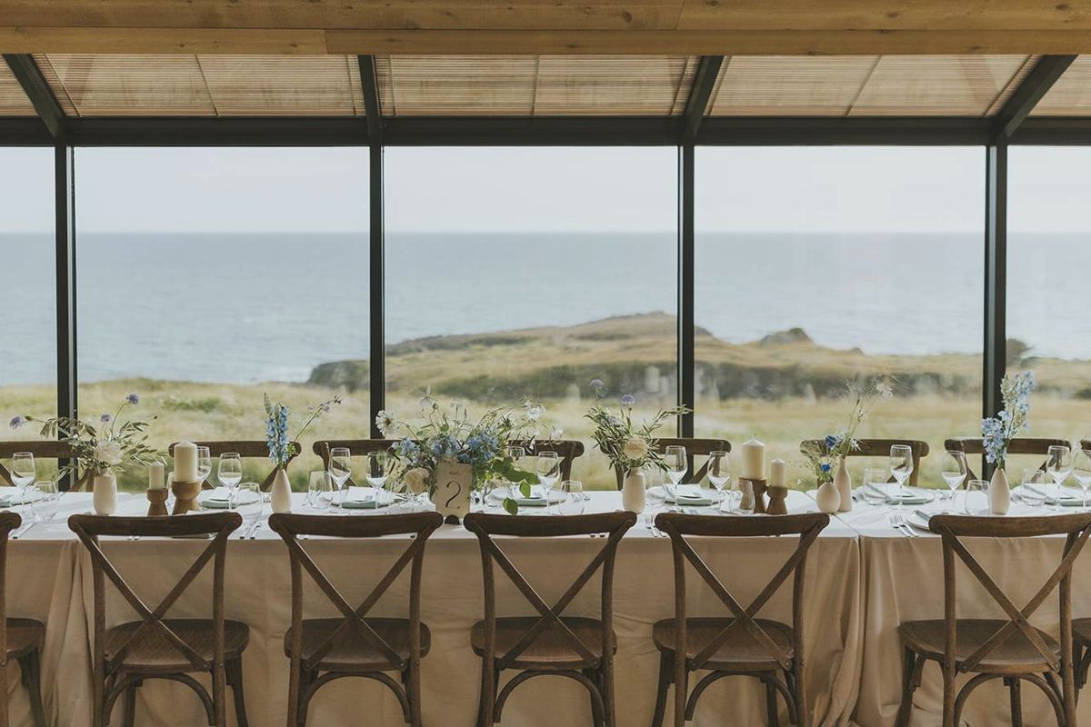 A long dinner table set in the glass-walled Solarium with the ocean and bluffs through the windows